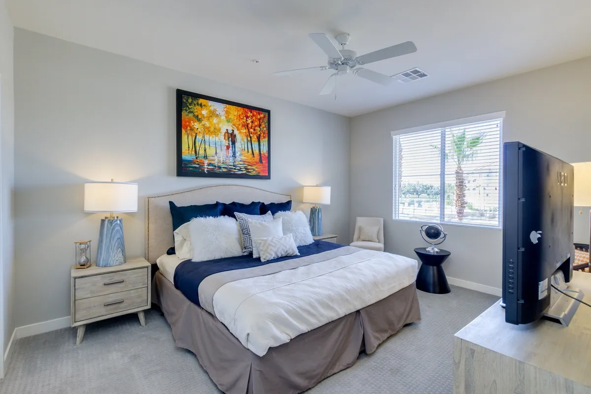 A serene primary bedroom suite is bathed in morning light, featuring a plush king bed with crisp linens, matching nightstands, tasteful artwork, a large window, and a personal TV situated perfectly for relaxing evenings. The overall atmosphere is cozy yet spacious, making it ideal for unwinding after a long day.
