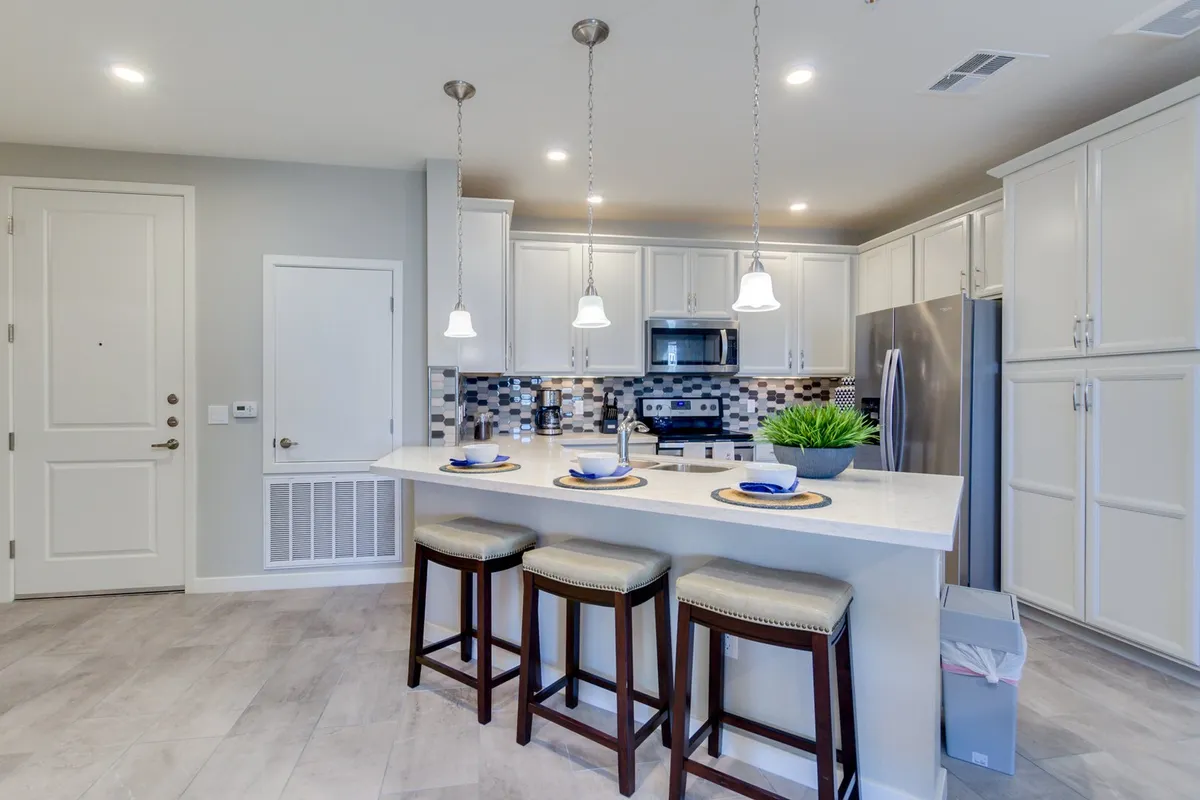 A wide, modern kitchen features a large island with pendant lighting, sleek stainless steel appliances, a colorful backsplash, and elegant dining settings. The open layout allows for seamless transition from the kitchen to the dining area, highlighting the quartz countertops and custom cabinetry.