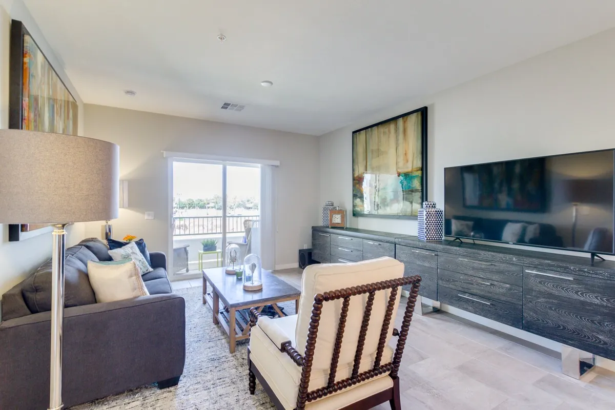 A plush seating area faces a large, wall-mounted television, with a coffee table and modern artwork completing the cozy living room setup. Sunlight filters through the window, emphasizing a bright and relaxing atmosphere.