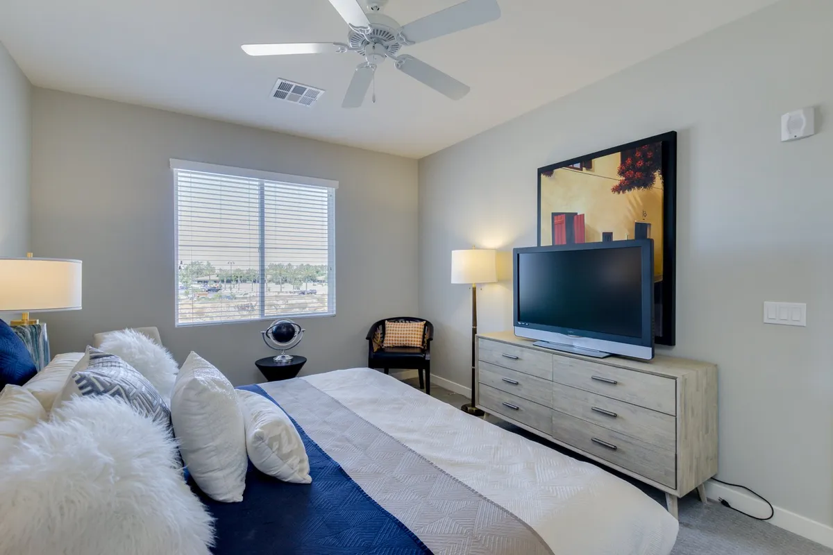 A comfortable second bedroom suite with a soft bed dressed in decorative pillows, a wall-mounted television, contemporary artwork, and a window that lets in gentle natural light. The space feels welcoming and is designed for restful sleep or a quiet spot to unwind.