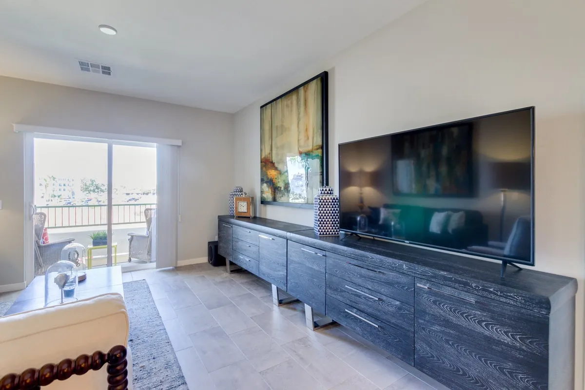 A spacious, sunlit living room outfitted for gatherings, featuring a large flat-screen television, an abstract painting as wall art, and a side table with decorative accessories. The overall mood is inviting, hinting at both lively social events and everyday relaxation.
